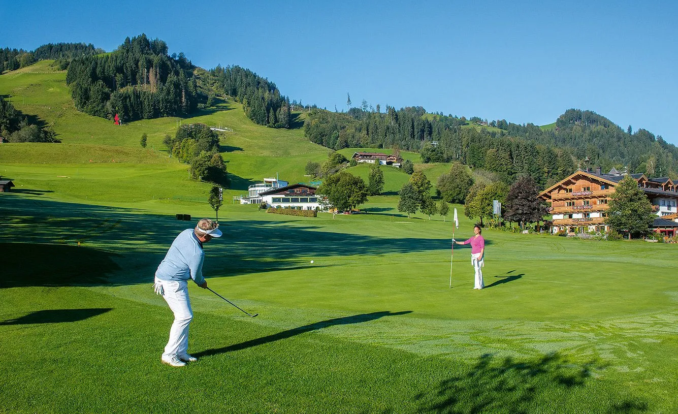 Golfer beim Chippen am Grün vor dem Hotel Rasmushof, dahinter bewaldete Hänge