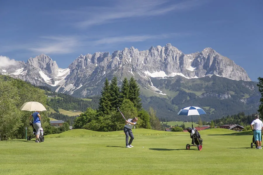 Golfer beim Abschlag vor dem Wilden Kaiser, daneben Trolleys mit Schirmen